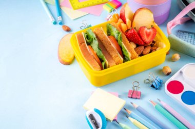 Okula dönelim. Sağlıklı okul yemekleri, çocuklar beslenme çantasına meyve, böğürtlen, fındık ve sebzeli sandviç koydu. Çocukların diyet yemekleri, eğitim okulu malzemeleri, parlak mavi arka plan manzarası.