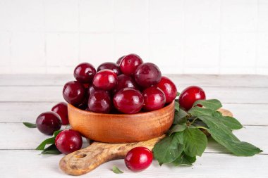 Yapraklı taze taze kiraz eriği. Bir kase lezzetli çiftlik, parlak kırmızı kirazlı erik meyveleri, ahşap çanak içinde beyaz ahşap arka planda fotokopi alanı.