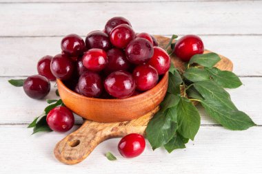 Yapraklı taze taze kiraz eriği. Bir kase lezzetli çiftlik, parlak kırmızı kirazlı erik meyveleri, ahşap çanak içinde beyaz ahşap arka planda fotokopi alanı.