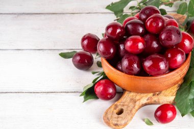 Yapraklı taze taze kiraz eriği. Bir kase lezzetli çiftlik, parlak kırmızı kirazlı erik meyveleri, ahşap çanak içinde beyaz ahşap arka planda fotokopi alanı.