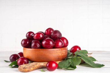 Yapraklı taze taze kiraz eriği. Bir kase lezzetli çiftlik, parlak kırmızı kirazlı erik meyveleri, ahşap çanak içinde beyaz ahşap arka planda fotokopi alanı.