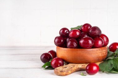 Yapraklı taze taze kiraz eriği. Bir kase lezzetli çiftlik, parlak kırmızı kirazlı erik meyveleri, ahşap çanak içinde beyaz ahşap arka planda fotokopi alanı.