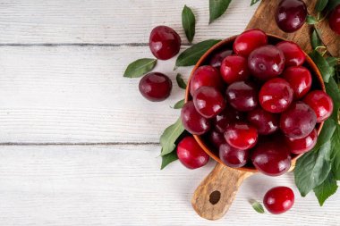 Yapraklı taze taze kiraz eriği. Bir kase lezzetli çiftlik, parlak kırmızı kirazlı erik meyveleri, ahşap çanak içinde beyaz ahşap arka planda fotokopi alanı.