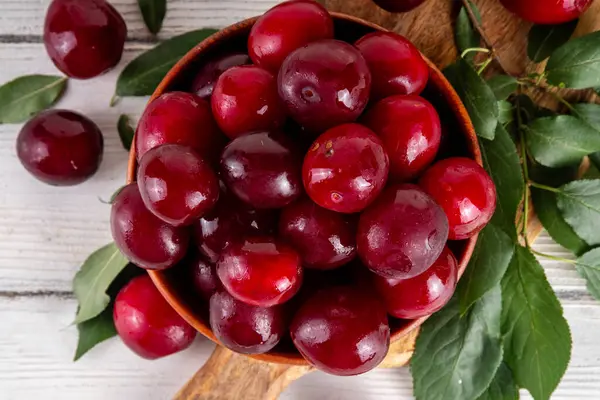 Yapraklı taze taze kiraz eriği. Bir kase lezzetli çiftlik, parlak kırmızı kirazlı erik meyveleri, ahşap çanak içinde beyaz ahşap arka planda fotokopi alanı.