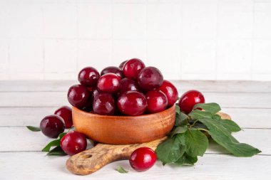 Yapraklı taze taze kiraz eriği. Bir kase lezzetli çiftlik, parlak kırmızı kirazlı erik meyveleri, ahşap çanak içinde beyaz ahşap arka planda fotokopi alanı.