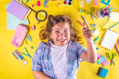 Sevimli duygusal kız okula dönmeye hazır çeşitli okul malzemeleriyle, parlak sarı arka planda eğitim aksesuarlarıyla.