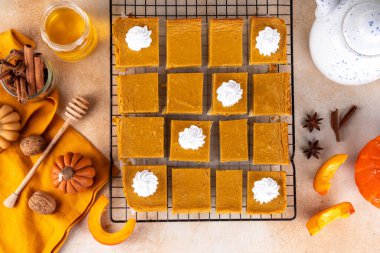 Homemade autumn taste pumpkin pie bars. Freshly baked pumpkin bars with whipped cream on it, with autumn decor and spices on beige table background, copy space 