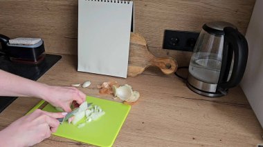 Mutfak ortamında, arka planda çaydanlık ve not defteri olan parlak yeşil bir kesme tahtasında soğan doğrayan kişi.