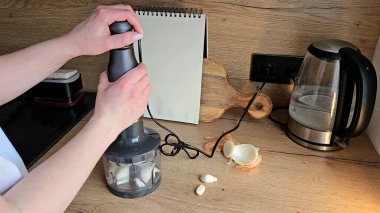 Çağdaş bir mutfak ortamında sarımsak presi kullanan eller taze sarımsak karanfilleri işliyor. Bir defter, kesme tahtası