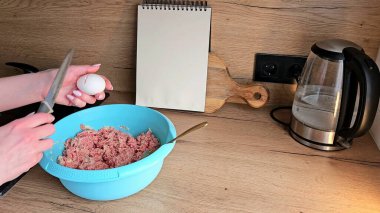 Elleri bir kase kıymalı ete yumurta kırmak üzere. Mutfakta, ahşap bir arka planda.. 