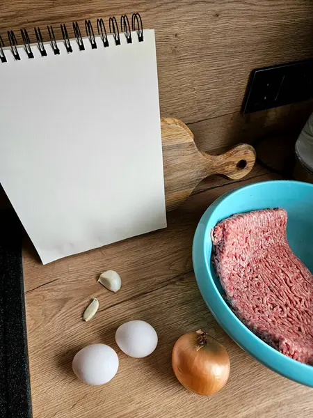 Yemek tarifi için gereken malzemeler arasında açık bir defter, kasede kıyma, iki yumurta, bütün bir soğan ve sarımsaklı karanfil bulunur..