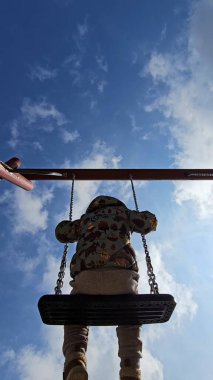 Çocuk salıncakta oturuyor, kabarık bulutlu açık mavi bir gökyüzüne bakıyor. Görüntü, çocukluk sevincini ve açık hava oyununu bir parkta yakalıyor.