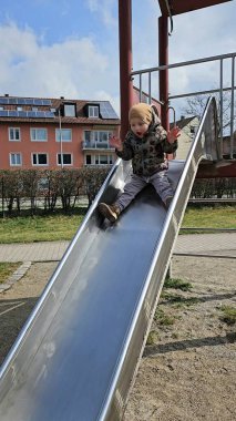 Metal bir oyun parkından kayan çocuk sıcak bir ceket ve bereyle meskun binalarla çevrili bir şekilde neşeyle kayıyor. Yüzlerinde heyecan ve mutluluğun ifadesi