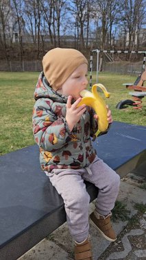 Sıcak kış kıyafetleri içinde çocuk oyun parkındaki bankta otururken muz yiyor. Çocuk çıplak ağaçlarla çevrili açık havada rahat ve mutlu görünüyor.