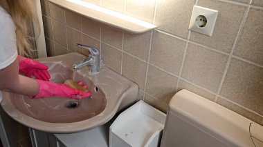 Pembe lastik eldiven takan eller akan suyun altında sünger kullanarak banyo lavabosunu temizliyor. Bej kaplama arkaplan ve musluk görünür, temizleme görevleri vurgulanır