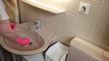 Elleri parlak pembe eldivenlerle banyo lavabosunu sünger ve temizlik malzemesi ile temizliyor. Odak noktası fayanslı banyo ortamında temizlik ve hijyen.