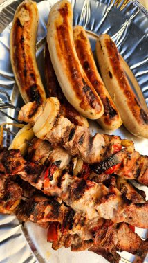 Izgara sosislerle dolu bir tabağın yakın çekimi ve yanık lekeler ve canlı renkler içeren lezzetli kebap şişleri. Barbekü ve açık hava aşçıları için mükemmel.