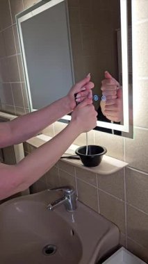 Elleri, banyo aynasının önünde bir kaseye boya sıkıyor. Kurulum saç rengi araçlarını içeriyor, bir ev ortamında boya uygulamasına hazırlık sürecini vurguluyor