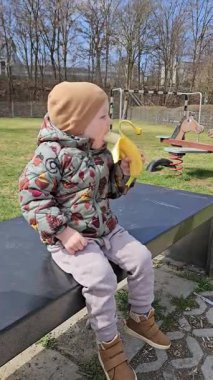 Renkli bir ceket giymiş genç bir çocuk elinde muz olan bir bankta oturuyor. Çocuk arka planda oyun ekipmanıyla dışarıda yemek yerken meraklı ve memnun görünüyor.