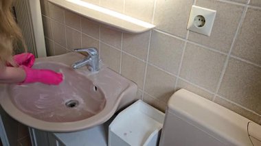Pembe eldiven giyen biri banyo lavabosunu süngerle temizler. Eylem, ev banyosundaki temizlik, temizlik ve bakımı yansıtıyor.