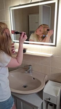 Kadınlar saç boyasını banyo aynasının önüne fırçayla sürerler. İstenilen saç rengine odaklanırlar. Sahne modern banyo düzenini ve kişisel bakım rutinini gösteriyor.