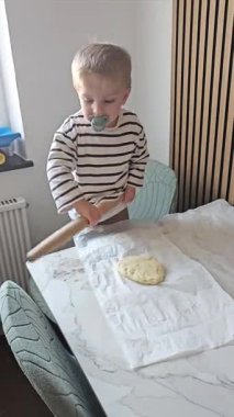 Beyaz bir yüzeyde tahta bir oklava ile hamuru yuvarlamaya odaklanan küçük bir çocuk. Çocuk bir mutfak ortamında odaklanma ve öğrenme gösteriyor