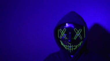Parlayan yeşil neon bir maske takan kapüşonlu bir şahıs gölgelerin arasında hareket ediyor, etrafında kırmızı ve mavi flaş ışıkları yanıp sönüyor. Titreşen parıltı, belirsizliği yoğunlaştırır, ürpertici bir ton oluşturur.