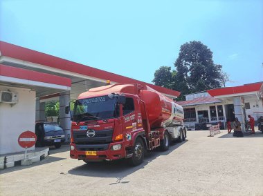 Bogor, Endonezya - 28 Eylül 2024: Bir benzin istasyonunda kırmızı bir Pertamina yakıt kamyonu, istasyonun kırmızı aksanları mavi gökyüzüne karşı dururken güneş ışığı altında yakıt teslimine hazır..