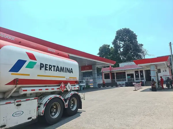 stock image Bogor, Indonesia - 28 September 2024: A Pertamina fuel truck delivers supply to a gas station on a sunny day, with the bright red signage of the station contrasting against the greenery in the background.