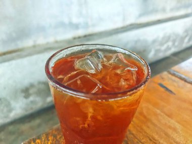 Kehribar rengi mükemmel olan ferahlatıcı bir bardak buzlu çay, kırsal ahşap bir masada servis edilir, sıcak bir günde serinlemek için ideal..