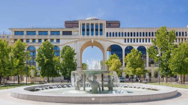 Berrak bir mavi gökyüzü altında yemyeşil ve modern mimarinin ortasında çarpıcı bir çeşme, güzel bir manzara yaratıyor.