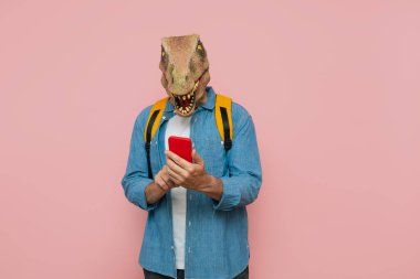 Sırt çantası ve pembe arka planda kertenkele maskesi olan adam akıllı telefonu kullanıyor..