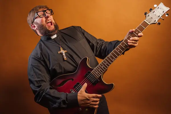 Portrait of a priest with a crucifix and black shirt playing the electric guitar.