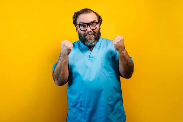 stock image The image depicts a bearded man wearing glasses, dressed in a light blue medical uniform, with clenched fists and a serious expression. The man is standing against a yellow background. The contrast between the vibrant yellow background and the light 