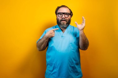 Mavi bluzlu ve gözlüklü bir adam sarı bir arka planın önünde duruyor, sağ eli havada. Küçük boyutu gösterir.