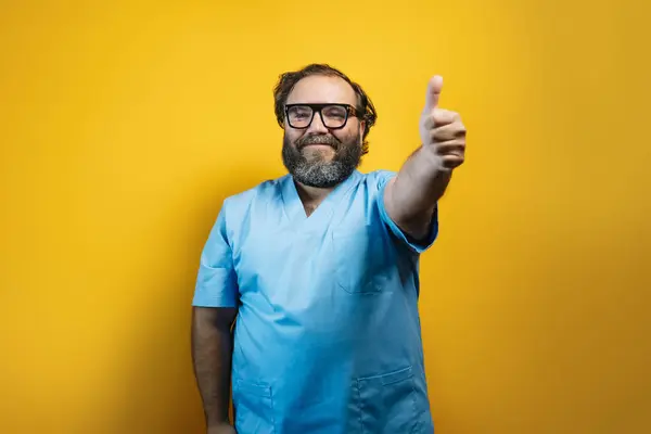 stock image Doctor in blue coat on yellow background makes a positive thumbs up gesture.