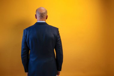 Görüntü koyu mavi takım elbiseli ve açık mavi gömlekli bir adamı gösteriyor. Sırtı sarı bir duvarın önünde kameraya dönük duruyor..