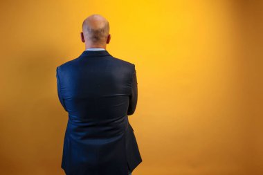 Görüntü koyu mavi takım elbiseli ve açık mavi gömlekli bir adamı gösteriyor. Sırtı sarı bir duvarın önünde kameraya dönük duruyor..