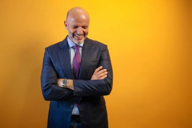 Sarı bir duvarın önünde duran mavi takım elbiseli ve açık mavi gömlekli bir adam gülümsüyor ve kollarını kavuşturuyor..