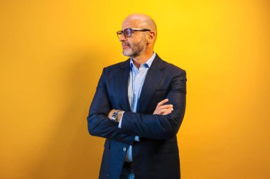 Koyu mavi takım elbiseli ve gözlüklü bir adam sarı bir duvarın önünde duruyor..