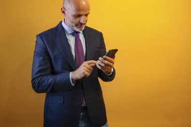 Mavi takım elbiseli, akıllı telefonunu kullanan bir adam sarı duvarın önünde duruyor..