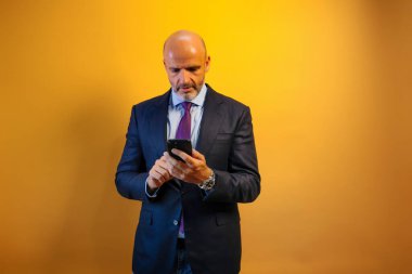 Mavi takım elbiseli, mavi gömlekli ve kot pantolonlu bir adam sol eli cebinde telefonuna bakıyor. Sarı bir duvarın önünde duruyor..