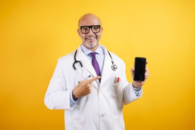 Beyaz önlüklü, dost canlısı bir doktor elinde akıllı bir telefon tutuyor. Modern sağlık ve iletişimi öneriyor..