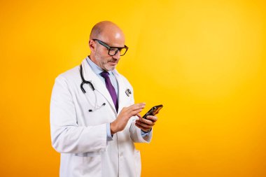 Beyaz önlüklü, dost canlısı bir doktor elinde akıllı bir telefon tutuyor. Modern sağlık ve iletişimi öneriyor..
