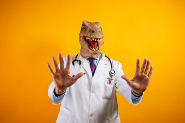 Sarı arka planda dinozor maskeli bir doktor. Avuç içini gösterir.