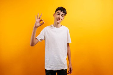 Genç çocuk gülümser, el hareketi yapar, sarı arka plan.