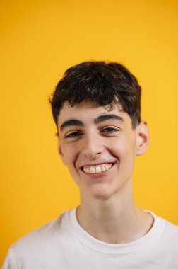 Beyaz tişörtlü, sarı arka planda gülümseyen genç çocuk..