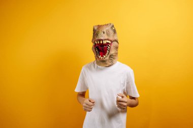 Sarı arka planda T-Rex maskeli bir genç.