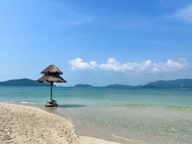 Sade ve tropikal bir plaj sahnesi, kumlu sahilde turkuvaz suyu ve berrak mavi gökyüzünün altındaki uzak dağlardan oluşan sazdan bir şemsiye.