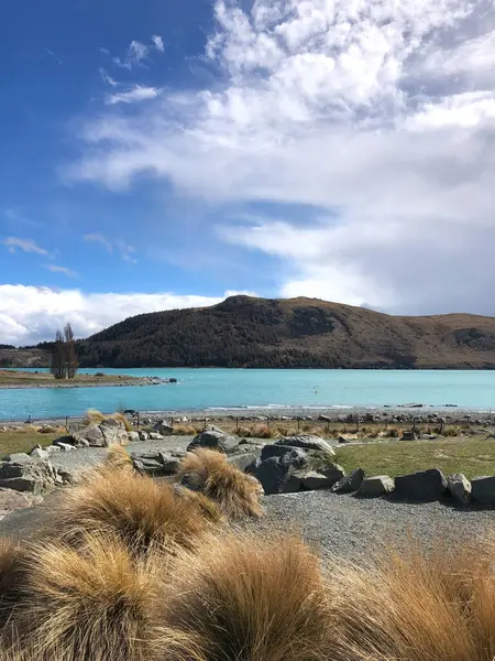 Parlak turkuaz bir göl manzarası. Etrafı altın otlar ve kayalık arazilerle çevrili. Arkaplanda dağlar ve bulutlarla dolu bir gökyüzü var.